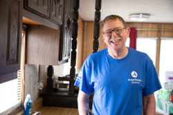 Photo showsa white man with glasses, short brown hair and a light mustache smiling at the camera. He is wearing a blue AmeriCorps Seniors t-shirt and standing in a kitchen with brown cupboards on the left frame of the photo.
