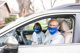 A white man and woman look at the camera from inside the front seats of a sedan. They are both wearing blue AmeriCorps face masks and gray  AmeriCorps logo t-shirts.. The woman has short, dark hair, and the man has no hair on his head and wears glasses.