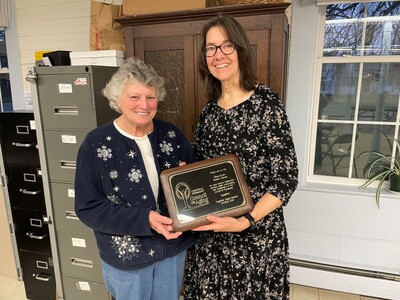 Photo of Phyllis Deering and Kathy Paquet. Phyllis stands on the left in front of a file cabinet. She has gray short hair and is wearing a blue cardigan over a white shirt and jeans. She holds half of a CVCOA Community of Strangth Plaque. Kathy holds the other half, on her right. She has brown shoulder-length hair, glasses, and a black dress with a light print all over it.